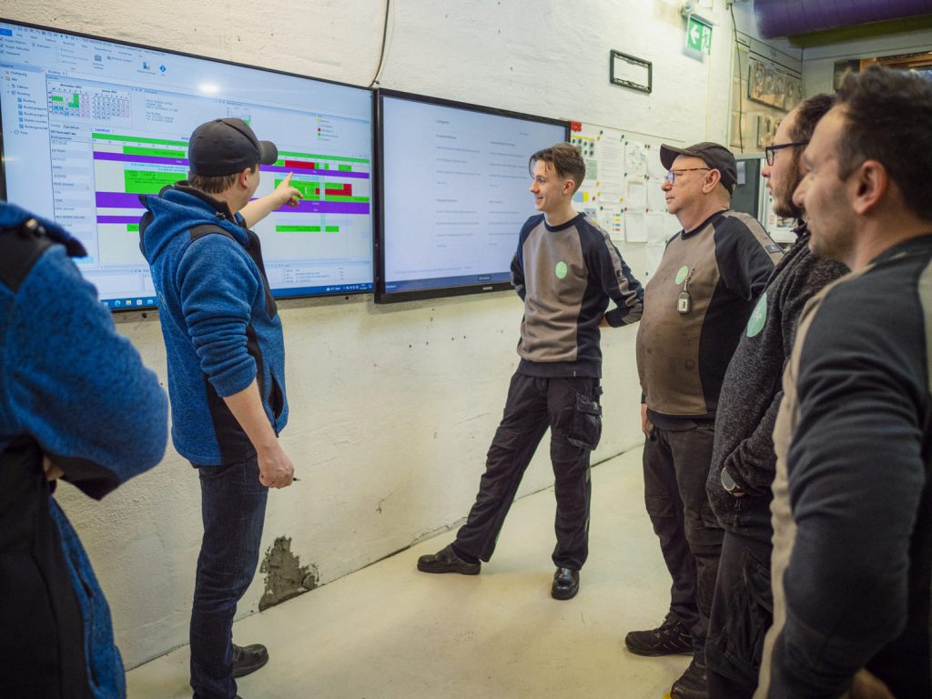 Fem personer som står foran en forbedrings tavle i et bilverksted. De diskuterer dagens oppgaver.