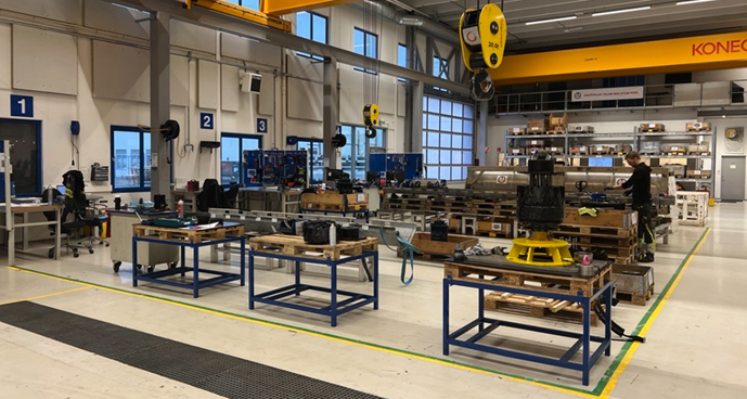 Et ryddig verkstedområde med arbeidsbenker og verktøy plassert systematisk, tydelig oppmerkede soner på gulvet og traverskran i taket, med maskindeler og utstyr organisert på pallereoler i bakgrunnen.